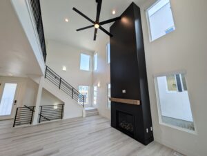 Spacious modern living room with vaulted ceiling, large black accent wall featuring a fireplace, and tall windows. A staircase with a black metal railing ascends to the left.