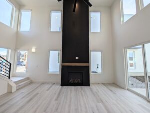 Spacious modern living room with high ceiling, black accent fireplace, large windows, light wood flooring, and a stairway on the left.