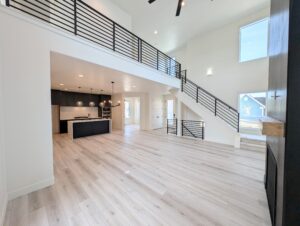 Spacious modern living room with high ceilings and a mezzanine. Features light wood flooring, black metal railings, and an open kitchen with a large island and stylish pendant lights.