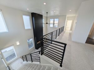 Spacious modern interior with a black metal railing, staircase, ceiling fan, and large windows. White walls and carpet create a bright, open atmosphere.