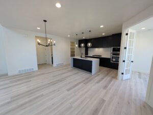 Modern kitchen and dining area with light wood flooring, black cabinetry, a central island, spherical pendant lights, and a black chandelier. Adjacent room visible through glass French doors.