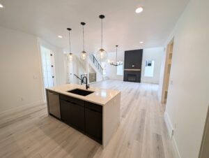 Modern open-concept kitchen and living area with a central island, pendant lights, light wood flooring, and a fireplace. Stairs visible in the background.