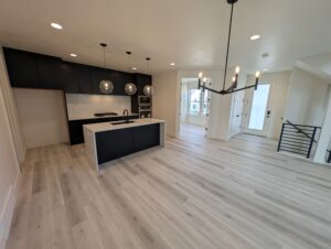 A modern kitchen with a white island, black cabinets, and pendant lights. Light wood flooring extends to an open living area with a chandelier. White walls and a doorway are visible.