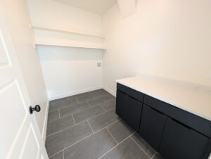 Minimalist laundry room with white walls, dark floor tiles, black cabinets, white countertop, and open shelving.