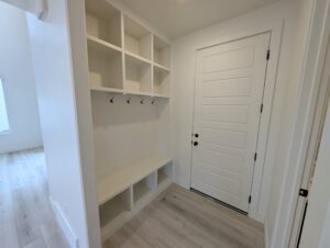 A small mudroom with white walls features a bench, open shelves, and hooks. There is a closed white door and light-colored wooden flooring.