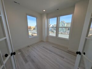 A bright, empty room with light wood flooring, white walls, and large windows overlooks a residential street. Two closed glass-paneled doors are visible at the entrance.