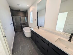 Modern bathroom with a double sink vanity, large mirror, standalone tub, and glass-enclosed shower. The design features dark tiles and black fixtures.