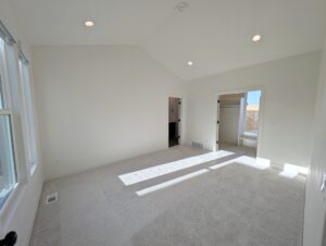 A bright, empty room with white walls and carpet, recessed lighting, and sunlight streaming through a large window. There are doorways leading to other spaces.