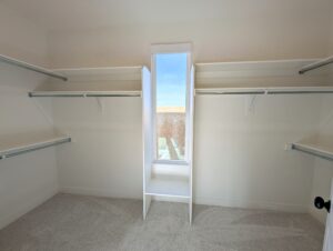 Empty walk-in closet with white walls, a small window in the center, and multiple horizontal shelves on both sides. Carpeted floor.