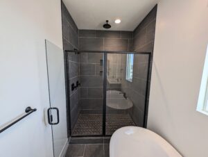 Modern bathroom with a glass-enclosed shower featuring dark tiles and a black showerhead. A white bathtub is visible in the foreground.