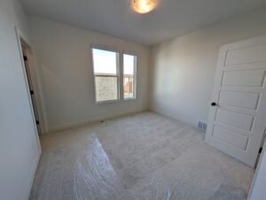 Empty room with beige carpet, white walls, and a ceiling light. There are two large windows and an open door. Plastic sheeting covers the floor.