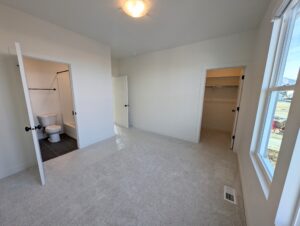 A vacant room with beige carpet, a window, an open door leading to a bathroom, and another to a closet.