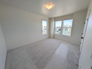 Empty room with white walls, carpeted floor, single ceiling light, and three large windows showing a neighborhood view.