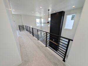 A modern interior with a high ceiling, black metal railing, large windows, and a ceiling fan. White walls and carpeted floor are visible.