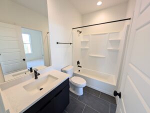 A modern bathroom with white walls, a bathtub with a shower, a toilet, a black vanity with a sink, and a mirror. The floor is tiled in gray.