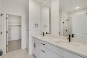 Modern bathroom with double sinks, large mirror, black fixtures, and a view into the Blue Jay #16 walk-in closet.