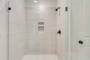 Modern glass-enclosed shower with white tile walls, a Blue Jay-inspired recessed shelf, and dark fixtures.