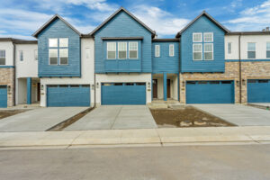 Row of modern townhouses with Blue Jay-hued facades, garage doors, and paved driveways under a cloudy sky.