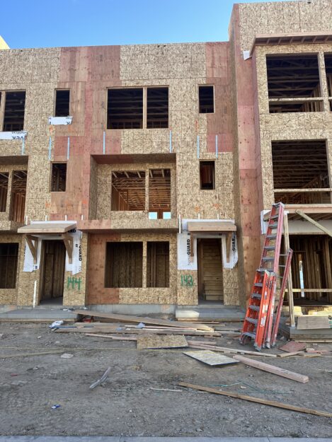 A multi-story building under construction with wooden framing, exposed OSB panels, and a red ladder leaning against the facade. Construction debris is scattered on the ground.