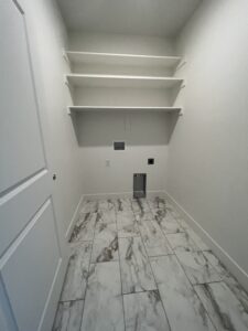 Empty laundry room with white walls, marble-patterned floor tiles, and three wall-mounted shelves. Washer and dryer hookups are visible on the back wall.