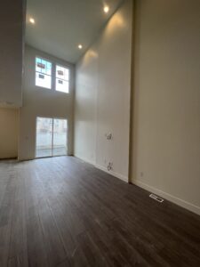 A spacious, empty room with high ceilings, dark wood flooring, and large windows. Walls are light colored, and there are visible electrical outlets on one side.