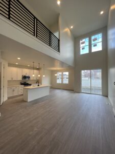 Bright open-concept living space with high ceilings, large windows, an upper loft, and a modern kitchen with an island and pendant lights.