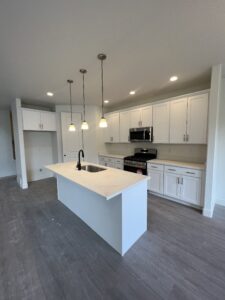 Modern kitchen with white cabinets, an island with a sink, hanging lights, and stainless steel appliances on hardwood floors.