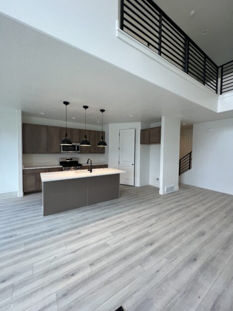 Open-concept kitchen and living area with light wood flooring, gray cabinets, black pendant lights, and a second-floor railing.