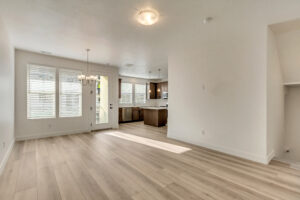 Bright, empty living space with light wood flooring, chandelier, and view into a modern kitchen with large windows.