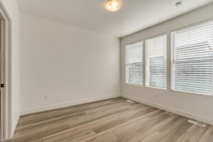 Empty room with wooden floors, white walls, and three large windows with blinds. Ceiling light fixture is visible.