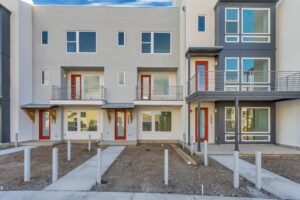 Modern townhouses with large windows, red doors, and balconies, adjacent to a bare dirt front yard with pathways.