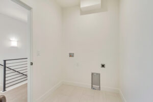 A small laundry area with white walls, a nightingale design on the detergent shelf, and an open door revealing a stairway with black railing.